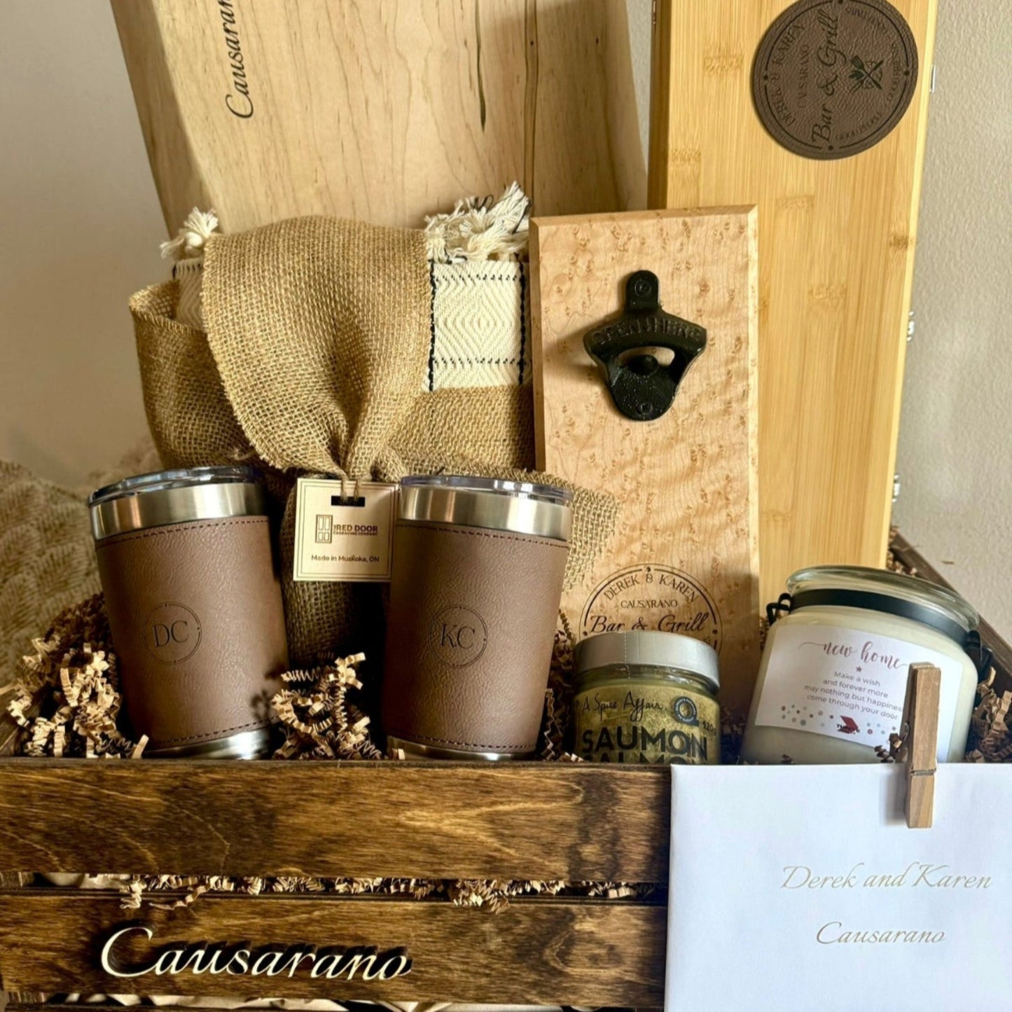 Rustic Luxe gift crate with personalized maple cutting board, bamboo BBQ tool set, wall bottle opener, custom candle, brown leatherette tumblers, Kana throw blanket, engraved family envelope, and crate with 3D family name cutout by The RDE Co.