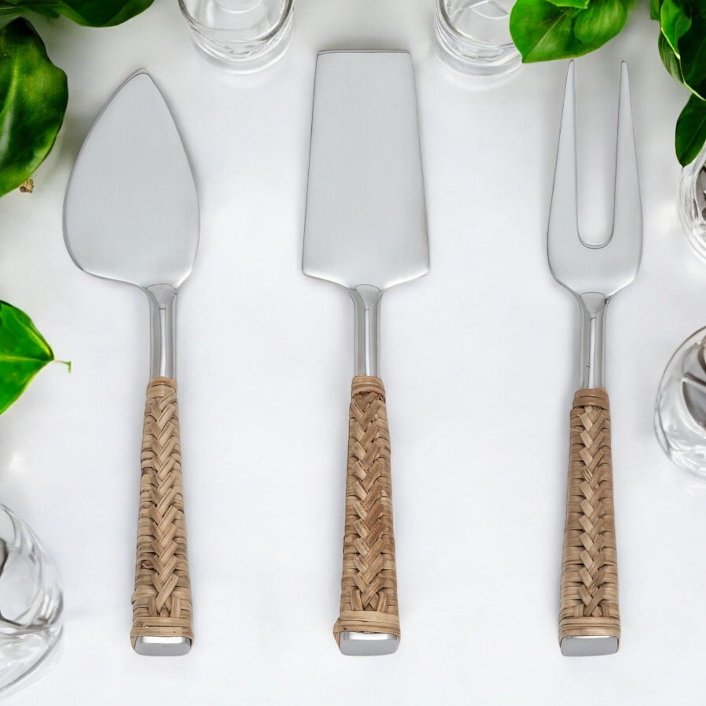 set of 3 stainless steel with rattan handle cheese knives on white background with glassware and greenery