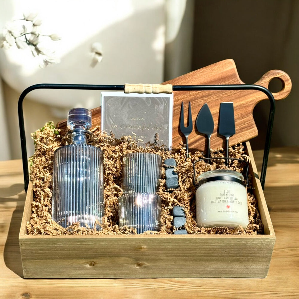 Gift set with decanter, tumbler glasses, scotch stones, cheese knives, and cheese board with a  candle in a wooden box on a table.