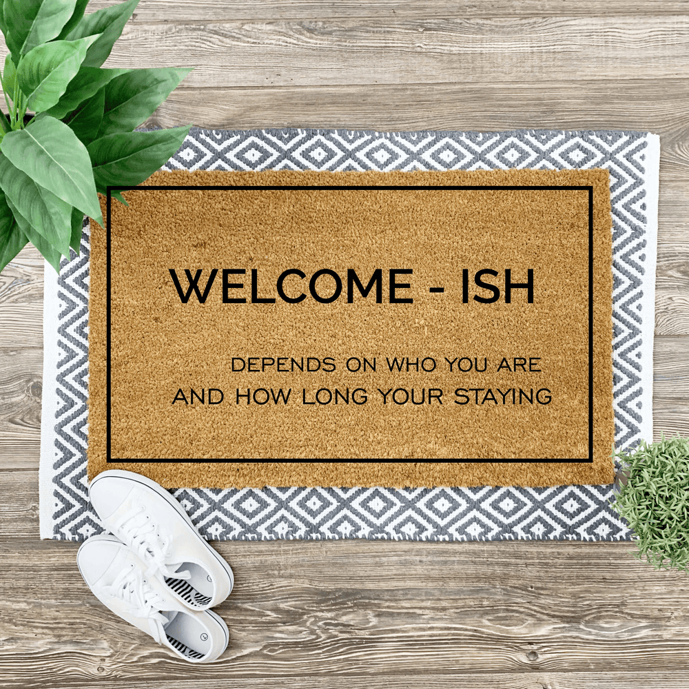 Coir door mat with funny saying on top of a woven rug with sneakers and a plant on the side