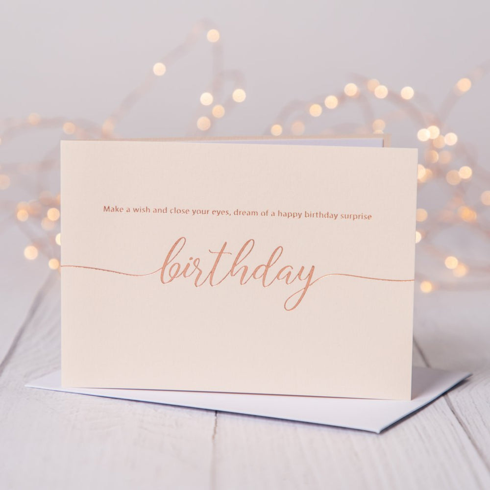 Blush pink birthday card with rose gold foil  saying Make a wish and close your eyes, dream of a happy birthday surprise. Card is on a table with fariy lights in the background