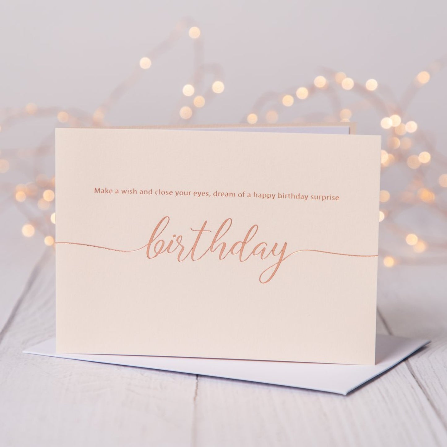 Blush pink birthday card with rose gold foil  saying Make a wish and close your eyes, dream of a happy birthday surprise. Card is on a table with fariy lights in the background