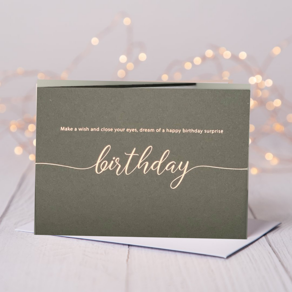 Grey luxe birthday card with rose gold foil  saying Make a wish and close your eyes, dream of a happy birthday surprise. Card is on a table with fariy lights in the background
